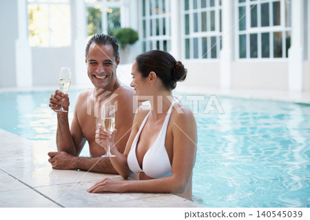 Couple, pool and drinking champagne on vacation together, romance and conversation at hotel. Happy people, summer holiday and love or bonding in water, laughing and glasses for alcohol on honeymoon 140545039