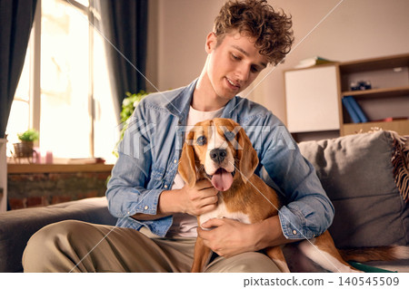 Young man hugging beagle on sofa in warm home interior with soft daylight. Young man hugging beagle on sofa in warm home interior with soft daylight. 140545509