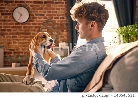 Young man relaxing with playful beagle dog on sofa in warm home interior. Young man relaxing with playful beagle dog on sofa in warm home interior. 140545531