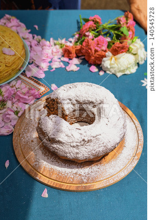 Bulgarian bundt cake with powdered sugar stands on wooden board with rose petals at Rose Festival in Kazanluk. Bread food display shows homemade baking, dessert tradition, festival food, local cuisine Bulgarian bundt cake with powdered sugar stands on wooden board with rose petals at Rose Festival in Kazanluk. Bread food display shows homemade baking, dessert tradition, festival food, local cuisine 140545728