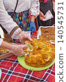 Pogacha bread at Bulgarian festival. Hands break pogacha bread on table with Bulgarian costumes, festival food, pastry, checked cloth. Pogacha bread shows cuisine, tradition, heritage, baking, event 140545731