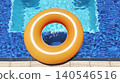 An orange inflatable swimming ring lies on the water of a hotel pool on a sunny summer day, with blue water visible through the hole in the ring. 140546516