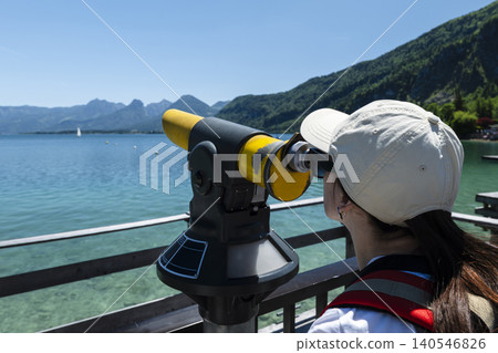 Woman looking through a coin operated telescope at mountains and lake Wolfgangsee in Austria, emphasizing outdoor observation Woman looking through a coin operated telescope at mountains and lake Wolfgangsee in Austria, emphasizing outdoor observation 140546826
