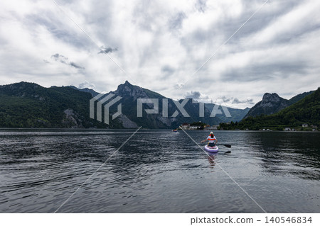 Paddleboarder moving on lake Traunsee with mountains and a lakeside settlement in the background, capturing a exploration moment. Paddleboarder moving on lake Traunsee with mountains and a lakeside settlement in the background, capturing a exploration moment. 140546834