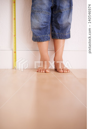 Child, wall and kid with tape measure for height, tracking growth or progress with feet in house. Legs, toddler and little person with development for size, length and stand on wooden floor at home 140546974