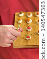 Hand of person taking canape from wooden board during reception, with canape bites and sauces behind on red tablecloth. Concept of catering service, tasting menu, appetizer tray, event hospitality 140547363