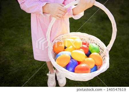 Child holding a white wicker basket filled with colorful plastic eggs in a grassy outdoor setting, wearing a pink gingham dress and white sneakers Child holding a white wicker basket filled with colorful plastic eggs in a grassy outdoor setting, wearing a pink gingham dress and white sneakers 140547619