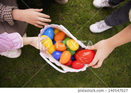 Children reaching for colorful plastic eggs from a white basket on green grass during an outdoor Easter egg hunt in a park setting Children reaching for colorful plastic eggs from a white basket on green grass during an outdoor Easter egg hunt in a park setting 140547627