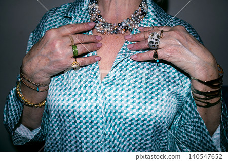 Woman wearing a patterned shirt showcases various jewelry pieces including rings, bracelets, and a pearl necklace, with hands positioned in front of her chest 140547652