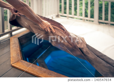 Person removing cover from a wooden hot tub revealing clear blue water on a deck surrounded by greenery, with a wooden railing in the background Person removing cover from a wooden hot tub revealing clear blue water on a deck surrounded by greenery, with a wooden railing in the background 140547654