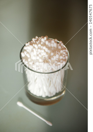 Glass container filled with cotton swabs on a smooth surface, one cotton swab placed beside the container, soft lighting enhances the clean appearance Glass container filled with cotton swabs on a smooth surface, one cotton swab placed beside the container, soft lighting enhances the clean appearance 140547675