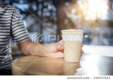 Hand of a person in a striped shirt holding a latte coffee in a clear cup on a wooden table with sunlight filtering through trees in the background Hand of a person in a striped shirt holding a latte coffee in a clear cup on a wooden table with sunlight filtering through trees in the background 140547683