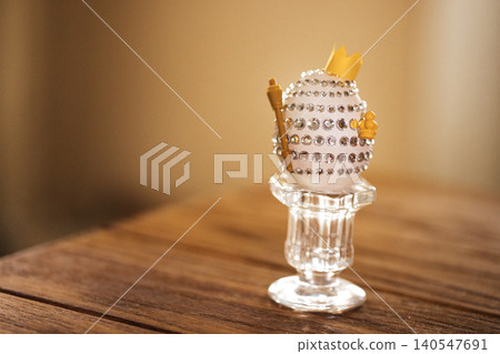 Decorative Easter egg adorned with rhinestones and a small crown, displayed on a crystal pedestal against a softly blurred background 140547691