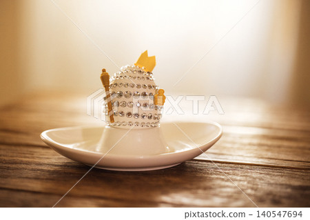 Decorative egg-shaped ornament adorned with rhinestones and a golden crown, placed on a white saucer atop a rustic wooden table with soft lighting in the background Decorative egg-shaped ornament adorned with rhinestones and a golden crown, placed on a white saucer atop a rustic wooden table with soft lighting in the background 140547694