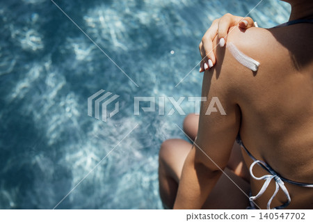 Female applying sunscreen on shoulder while sitting by the edge of a swimming pool with shimmering water reflecting sunlight in the background Female applying sunscreen on shoulder while sitting by the edge of a swimming pool with shimmering water reflecting sunlight in the background 140547702