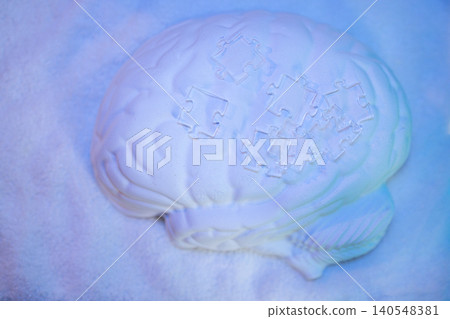 White brain sculpture with missing puzzle pieces on a textured surface illuminated by blue and purple lighting, creating a visually striking artistic display White brain sculpture with missing puzzle pieces on a textured surface illuminated by blue and purple lighting, creating a visually striking artistic display 140548381