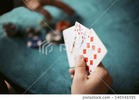 Hand holding playing cards with red diamonds and a colorful assortment of poker chips on a green felt table in a casual gaming setting Hand holding playing cards with red diamonds and a colorful assortment of poker chips on a green felt table in a casual gaming setting 140548395
