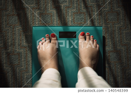 Female feet with red nail polish standing on a turquoise bathroom scale on a patterned carpet, sunlight casting shadows across the scene Female feet with red nail polish standing on a turquoise bathroom scale on a patterned carpet, sunlight casting shadows across the scene 140548399