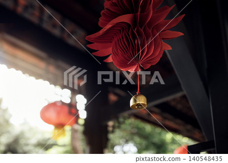 Red paper lantern decorations hanging from wooden beams in a traditional setting with blurred greenery and additional lanterns in the background 140548435