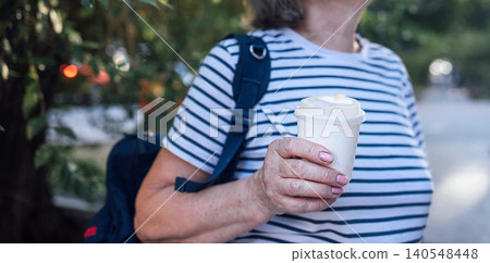 Senior woman wearing striped shirt holds a coffee cup while walking outdoors, with a backpack slung over her shoulder and greenery in the background Senior woman wearing striped shirt holds a coffee cup while walking outdoors, with a backpack slung over her shoulder and greenery in the background 140548448