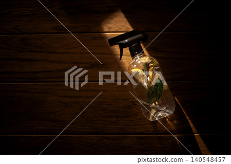 Transparent spray bottle with a black nozzle containing water and green leaves, illuminated by soft light on a wooden surface Transparent spray bottle with a black nozzle containing water and green leaves, illuminated by soft light on a wooden surface 140548457