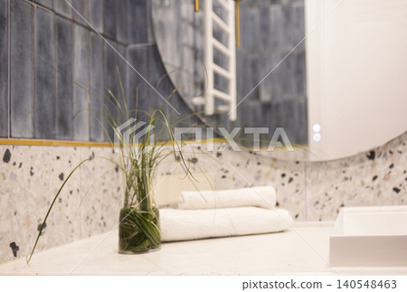 Modern bathroom interior featuring a round mirror, decorative plant in a glass vase, and neatly rolled white towels on a countertop with textured surfaces Modern bathroom interior featuring a round mirror, decorative plant in a glass vase, and neatly rolled white towels on a countertop with textured surfaces 140548463