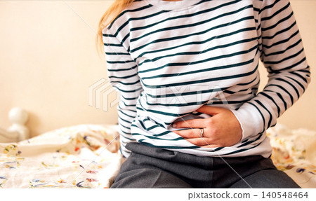 Female in striped long-sleeve shirt sitting on bed, holding stomach with discomfort, floral-patterned blanket and neutral wall in background Female in striped long-sleeve shirt sitting on bed, holding stomach with discomfort, floral-patterned blanket and neutral wall in background 140548464
