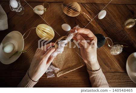Hands of a woman wrapping a small gift with decorative ribbon surrounded by Easter ornaments and eggs on a rustic wooden table Hands of a woman wrapping a small gift with decorative ribbon surrounded by Easter ornaments and eggs on a rustic wooden table 140548465