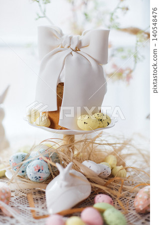 Easter cake decorated with a large white bow on a cake stand, surrounded by colorful eggs and decorative elements on a lace tablecloth Easter cake decorated with a large white bow on a cake stand, surrounded by colorful eggs and decorative elements on a lace tablecloth 140548476