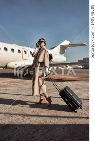 Stylish Caucasian mid adult woman talking on phone pulling suitcase on airport tarmac Stylish Caucasian mid adult woman talking on phone pulling suitcase on airport tarmac 140548689