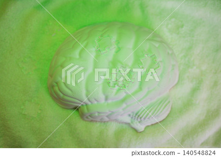 White brain-shaped model placed on a soft surface with green lighting, featuring puzzle piece designs embossed on the surface for educational or decorative purposes White brain-shaped model placed on a soft surface with green lighting, featuring puzzle piece designs embossed on the surface for educational or decorative purposes 140548824
