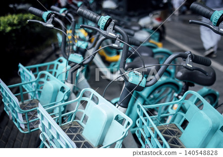 Multiple teal bicycles with front baskets parked in a row on a city street, showcasing handlebars and wheels, with blurred background of urban environment 140548828