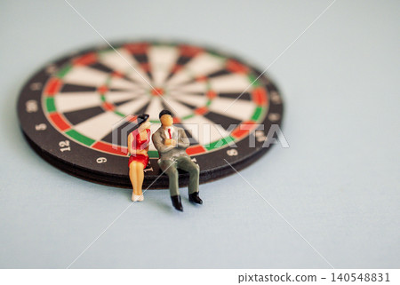 Miniature figurines of a man and woman sitting on a dartboard, with a soft blue background creating a playful and whimsical scene Miniature figurines of a man and woman sitting on a dartboard, with a soft blue background creating a playful and whimsical scene 140548831