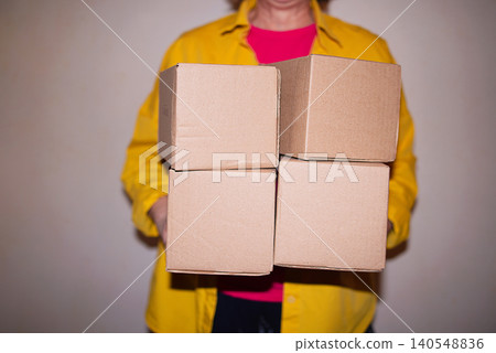 Individual holding four stacked cardboard boxes in front of a plain wall, wearing a yellow shirt over a pink top, showcasing a simple indoor setting Individual holding four stacked cardboard boxes in front of a plain wall, wearing a yellow shirt over a pink top, showcasing a simple indoor setting 140548836
