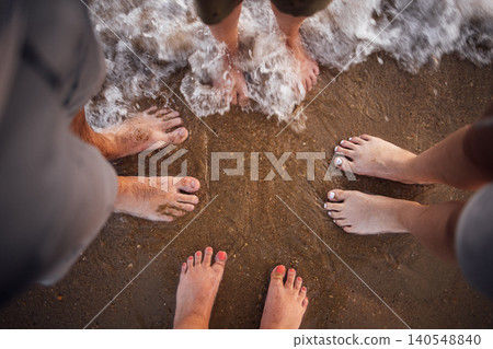 Group of four diverse feet standing on sandy beach shore with gentle waves lapping at toes during sunset, capturing a moment of relaxation and connection with nature Group of four diverse feet standing on sandy beach shore with gentle waves lapping at toes during sunset, capturing a moment of relaxation and connection with nature 140548840