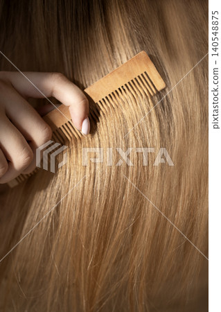 Close-up of a hand holding a wooden comb while brushing long, straight blonde hair, showcasing smooth strands and natural shine in soft lighting Close-up of a hand holding a wooden comb while brushing long, straight blonde hair, showcasing smooth strands and natural shine in soft lighting 140548875