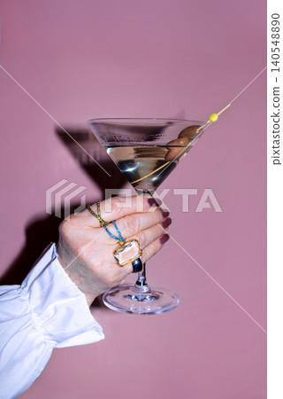 Hand holding a martini glass with an olive garnish against a pink background, showcasing elegant jewelry and manicured nails in a stylish presentation 140548890