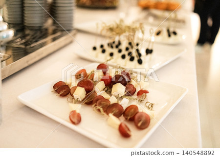 Assorted appetizers featuring skewered grapes and cheese cubes on white plates, elegantly arranged for a catering event with additional dishes in the background Assorted appetizers featuring skewered grapes and cheese cubes on white plates, elegantly arranged for a catering event with additional dishes in the background 140548892