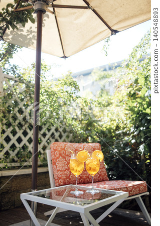 Two refreshing orange aperol spritz cocktails with slices of orange on a white table under a sun umbrella beside a cozy chair in a lush garden setting Two refreshing orange aperol spritz cocktails with slices of orange on a white table under a sun umbrella beside a cozy chair in a lush garden setting 140548893