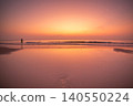 Lone figure stands in the ocean waves during a vibrant sunset with a crescent moon 140550224