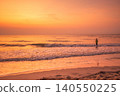 Lone figure wading into the ocean at sunrise with warm golden light reflecting on the water 140550225