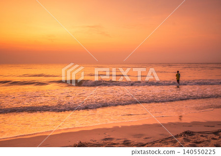 Lone figure wading into the ocean at sunrise with warm golden light reflecting on the water 140550225