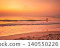 Lone child stands in the ocean waves during a vibrant orange sunset 140550226
