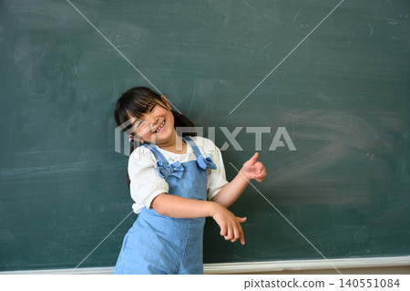 A female elementary school student smiles and points in front of the classroom blackboard. A female elementary school student smiles and points in front of the classroom blackboard. 140551084