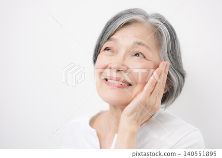 一張特寫鏡頭,一位頭髮花白的老婦人站在白色背景前,面帶微笑,輕撫著臉頰——這是一張美麗的照片。 一張特寫鏡頭,一位頭髮花白的老婦人站在白色背景前,面帶微笑,輕撫著臉頰——這是一張美麗的照片。 140551895