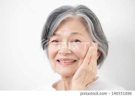 一張特寫美照，照片中一位頭髮花白的老婦人站在白色背景前，面帶微笑，直視鏡頭，用手輕撫臉頰。 140551896
