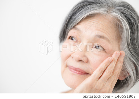 A close-up of an elderly woman (senior) with gray hair against a white background; she smiles and looks up, touching her cheek with her hand—a beauty image. 140551902