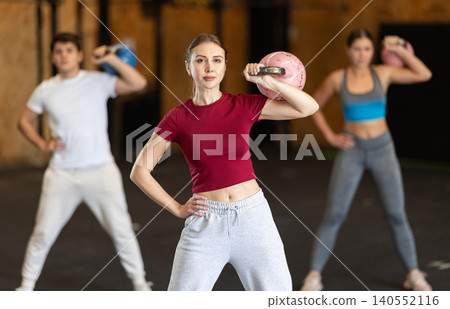 Girl in gym does exercises with kettlebell, lifts and lowers heavy kettlebell with one hand 140552116