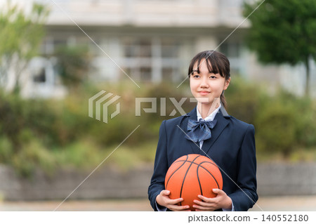 High school, junior high school, and elementary school girls in uniform standing in a schoolyard holding a basketball 140552180