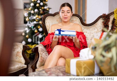 Excited young European woman in red dress enjoying wrapped gift while resting on couch before New Year holiday at flat Excited young European woman in red dress enjoying wrapped gift while resting on couch before New Year holiday at flat 140553492